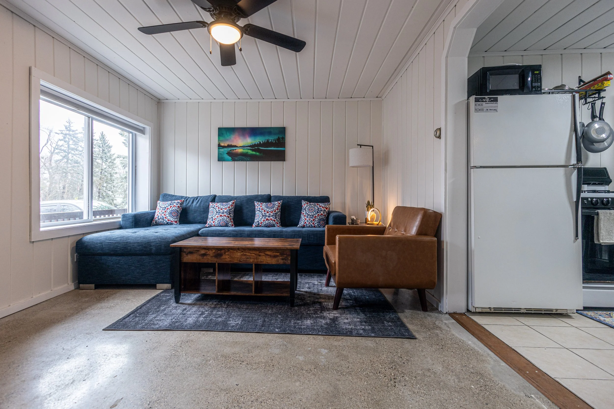 Living room with white paneled walls, large window, blue sofa with patterned pillows, wooden coffee table, brown leather armchair, floor lamp, wall art, ceiling fan, adjacent kitchen with white refrigerator, microwave, stove, and hanging pots.