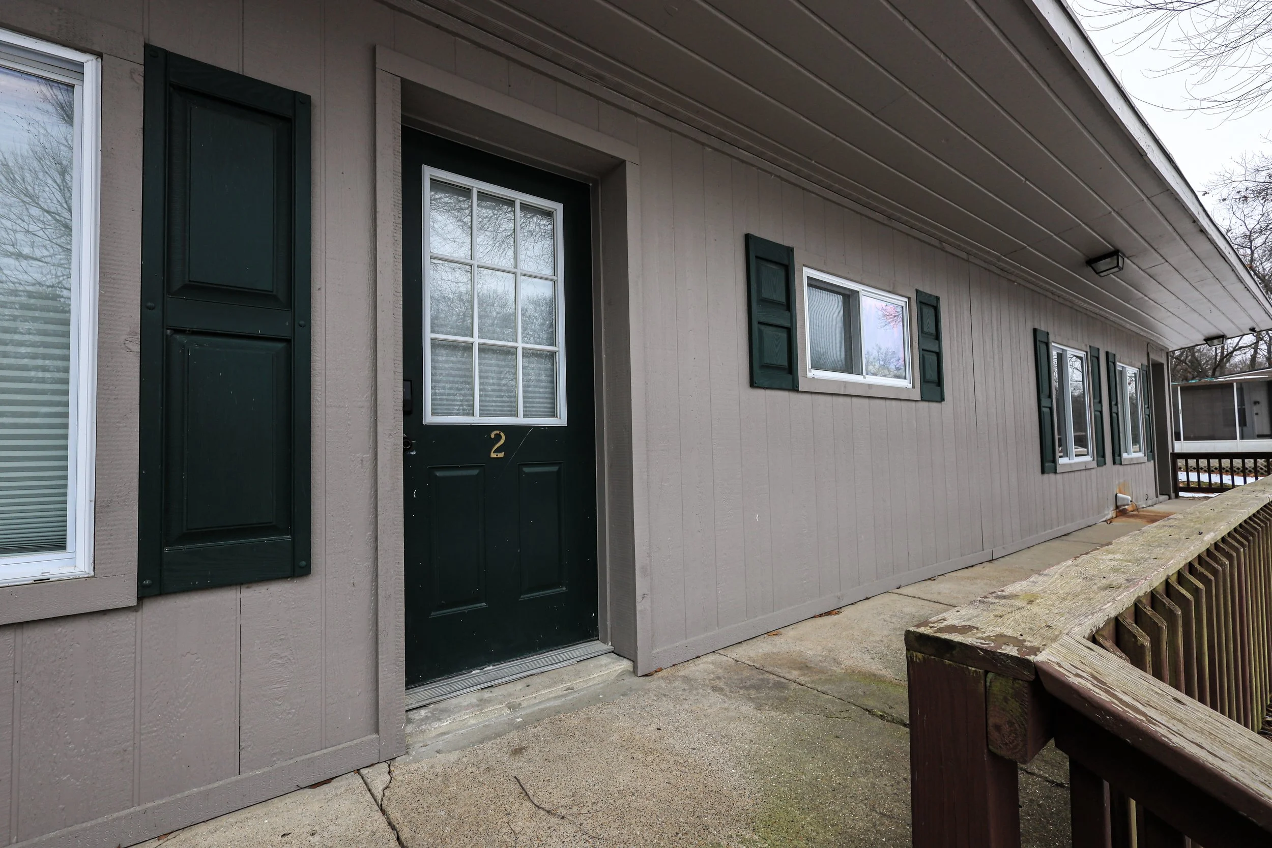 Exterior view of a beige apartment building with green shutters and a black door marked with the number 2, along with a concrete walkway and wooden railing.