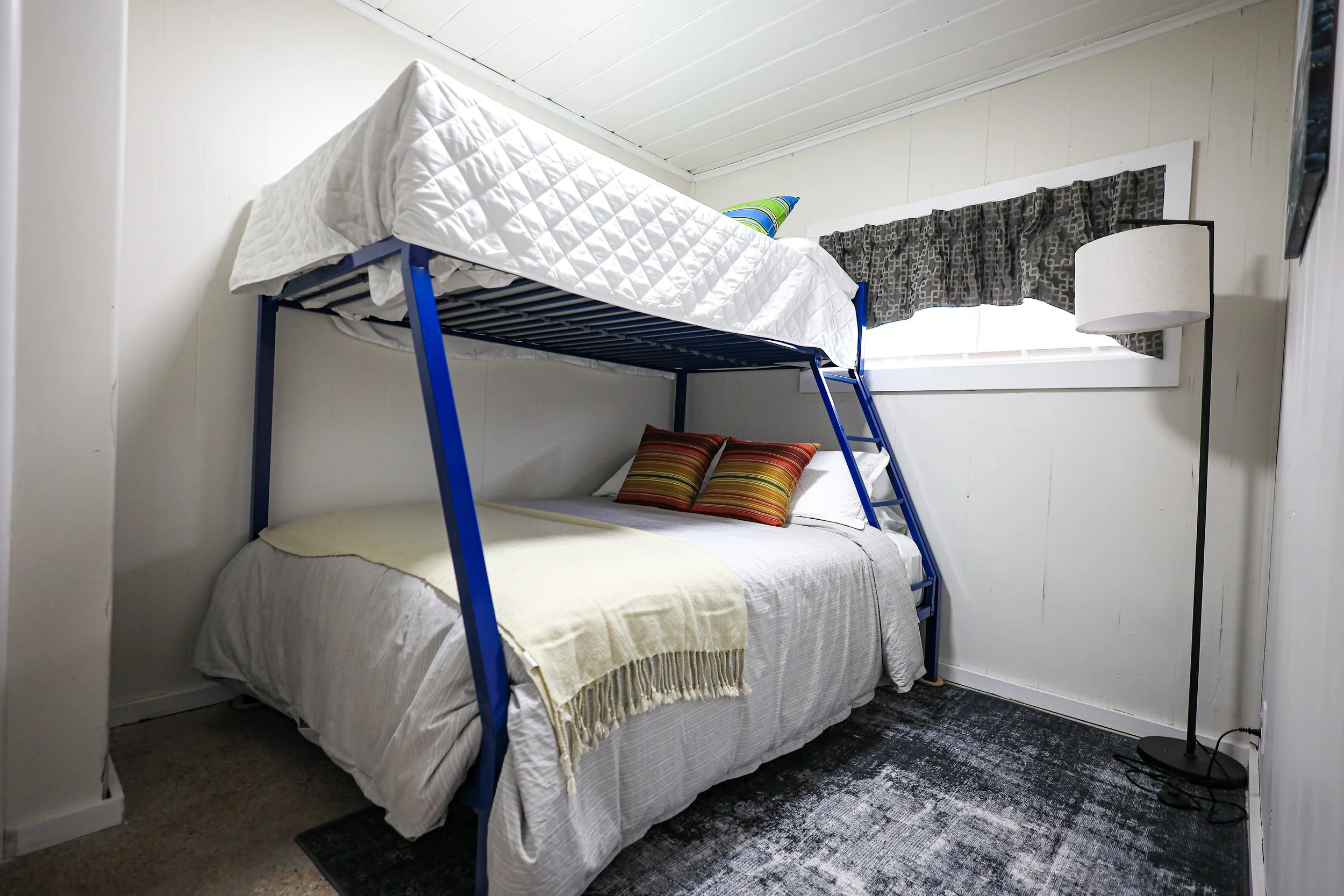 A bedroom with a bunk bed featuring a blue metal frame, with a lower full-size bed dressed in white sheets and two colorful striped pillows, and an upper twin-sized bed with a white quilted mattress, a green pillow, and a blanket. There's a small win