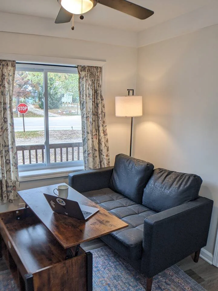 A cozy living room corner with a black loveseat, a wooden dining table with a laptop and a coffee mug, a standing floor lamp, a ceiling fan, and a window with floral curtains in The Nook showing a street view in Port Austin, MI.