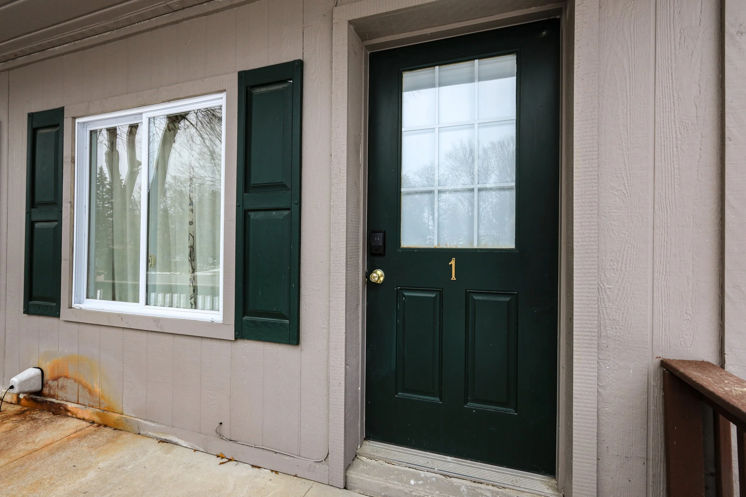 Front door with house number 1 and window, siding with green shutters, and a concrete porch with rust stains.
