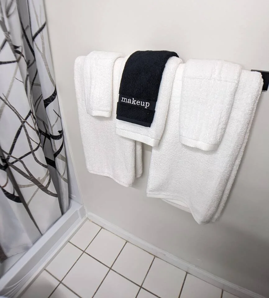 Three white towels, a black towel with 'makeup' embroidered on it, and another white towel hanging on a black towel rack in a bathroom, tiled floor, and a patterned shower curtain.