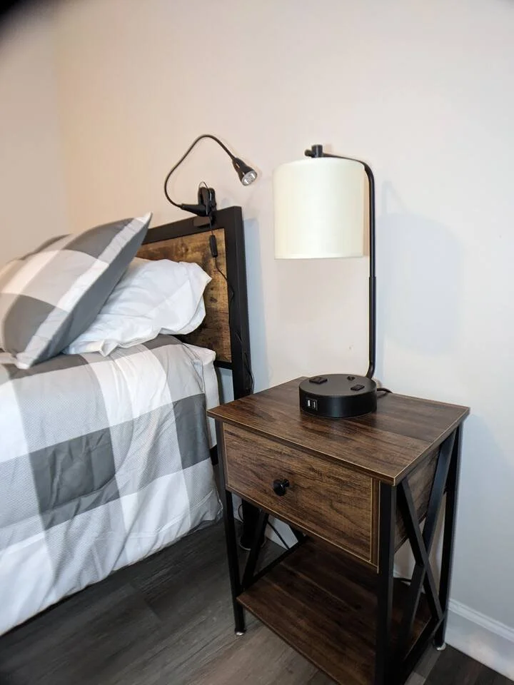 A bedside table with a drawer and a lower shelf next to a bed with plaid bedding, a headboard, and reading lamps mounted on the headboard and wall.