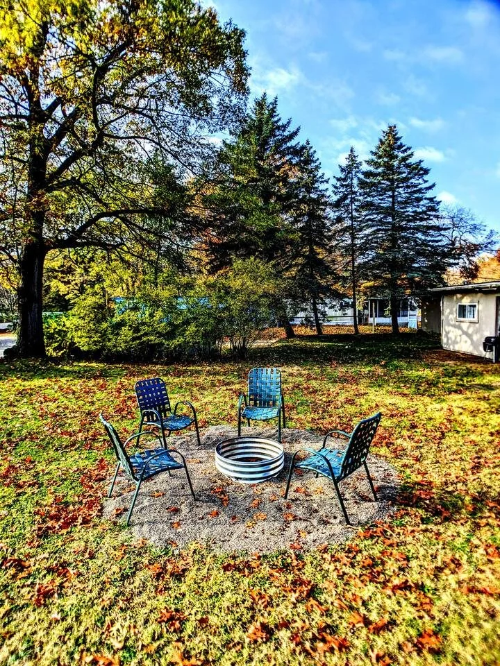 Four metal chairs arranged in a circle around a metal fire ring on a patch of dirt, with fallen leaves on grass and trees in the background under a partly cloudy sky.