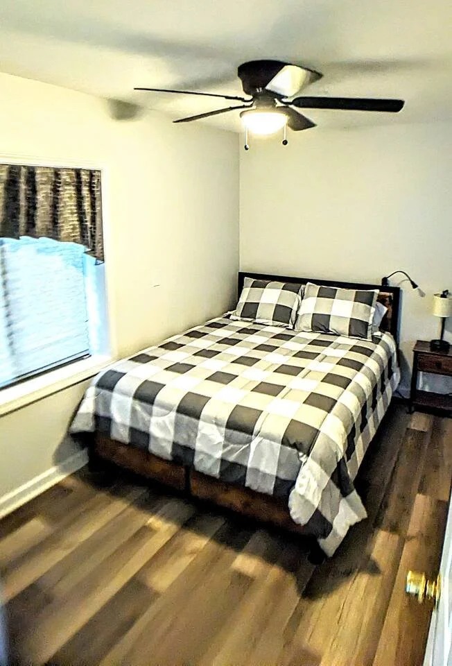 A bedroom with a bed that has a black and white checkered quilt and matching pillows, a window with patterned curtains, a wooden nightstand with a lamp, a ceiling fan with a light, and hardwood flooring.