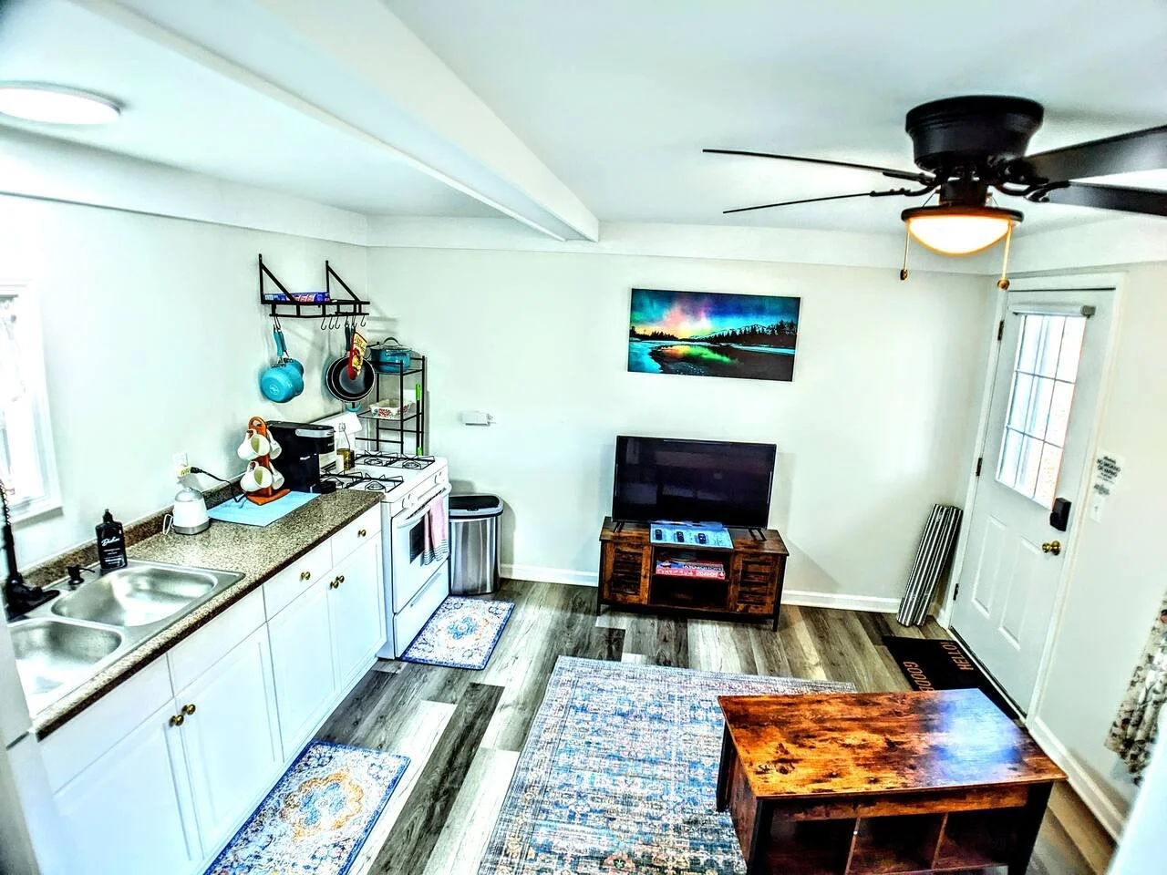 Living room and kitchen area with white cabinets, a small stove, a double sink, a TV on a wooden stand, and a ceiling fan with a light. There are rugs on the wood floor, and a picture of the northern lights on the wall.