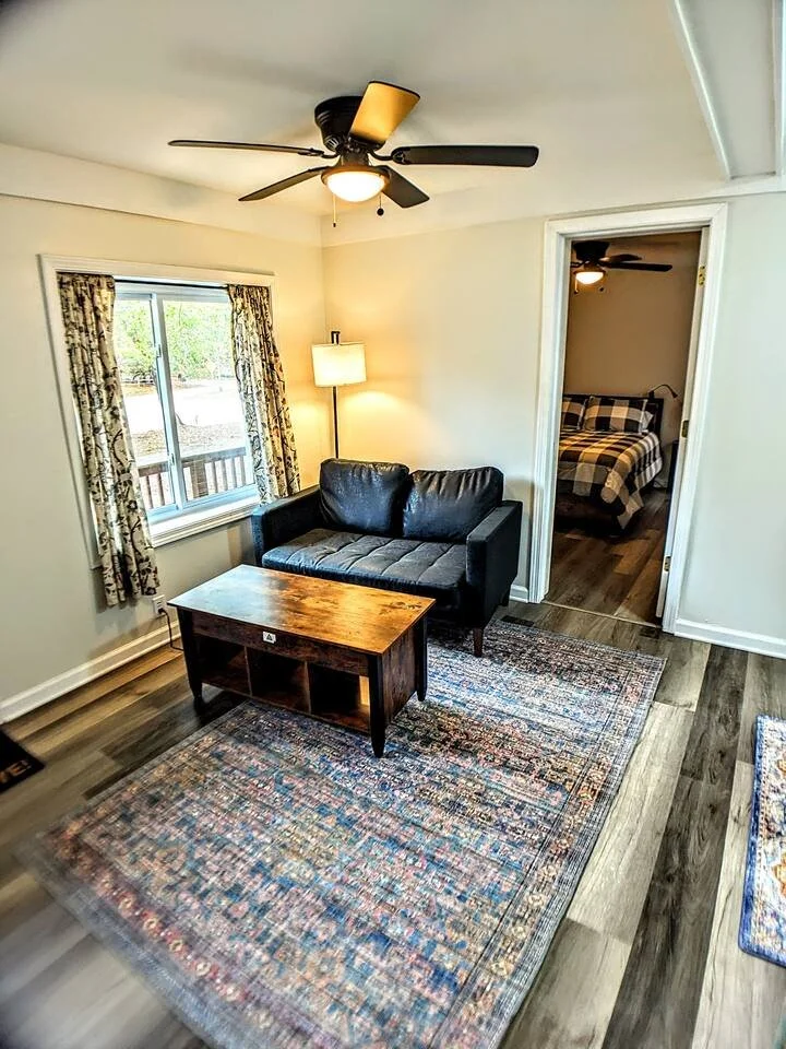 Living room with a black leather couch, wooden coffee table, colorful patterned area rug, window with floral curtains, floor lamp, ceiling fan with light, and doorway leading to a bedroom with a checkered bedspread and matching ceiling fan.