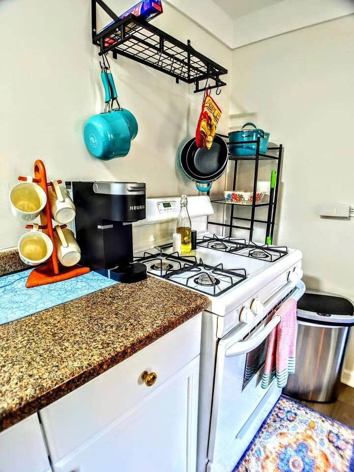 A small kitchen with a granite countertop, a white stove, and a black hanging rack with pots and pans. There is a coffee maker, a coffee mug holder with mugs, a soap dispenser, and a bottle of olive oil on the counter. A colorful rug is on the floor.
