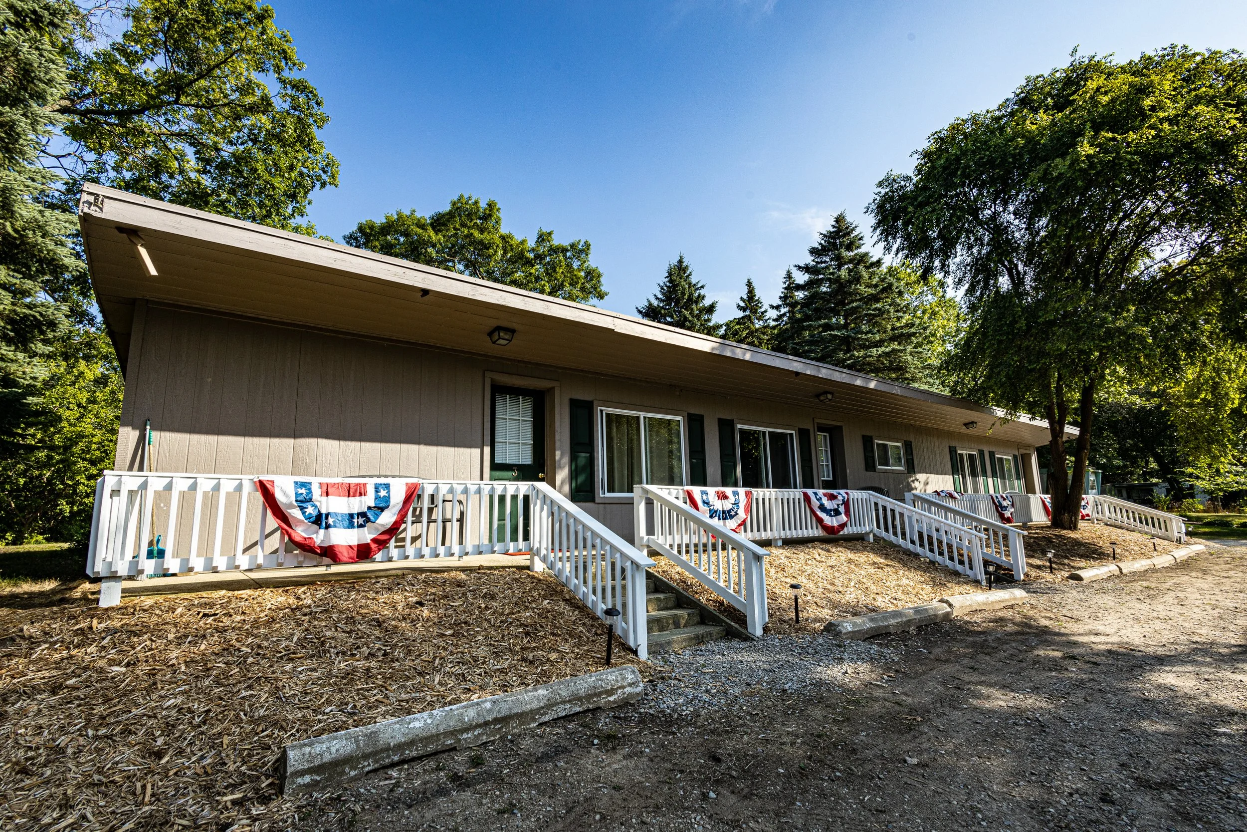 Single-story building called The Vintage with a beige exterior, white railings, and patriotic bunting decorations, surrounded by green trees and a clear blue sky.