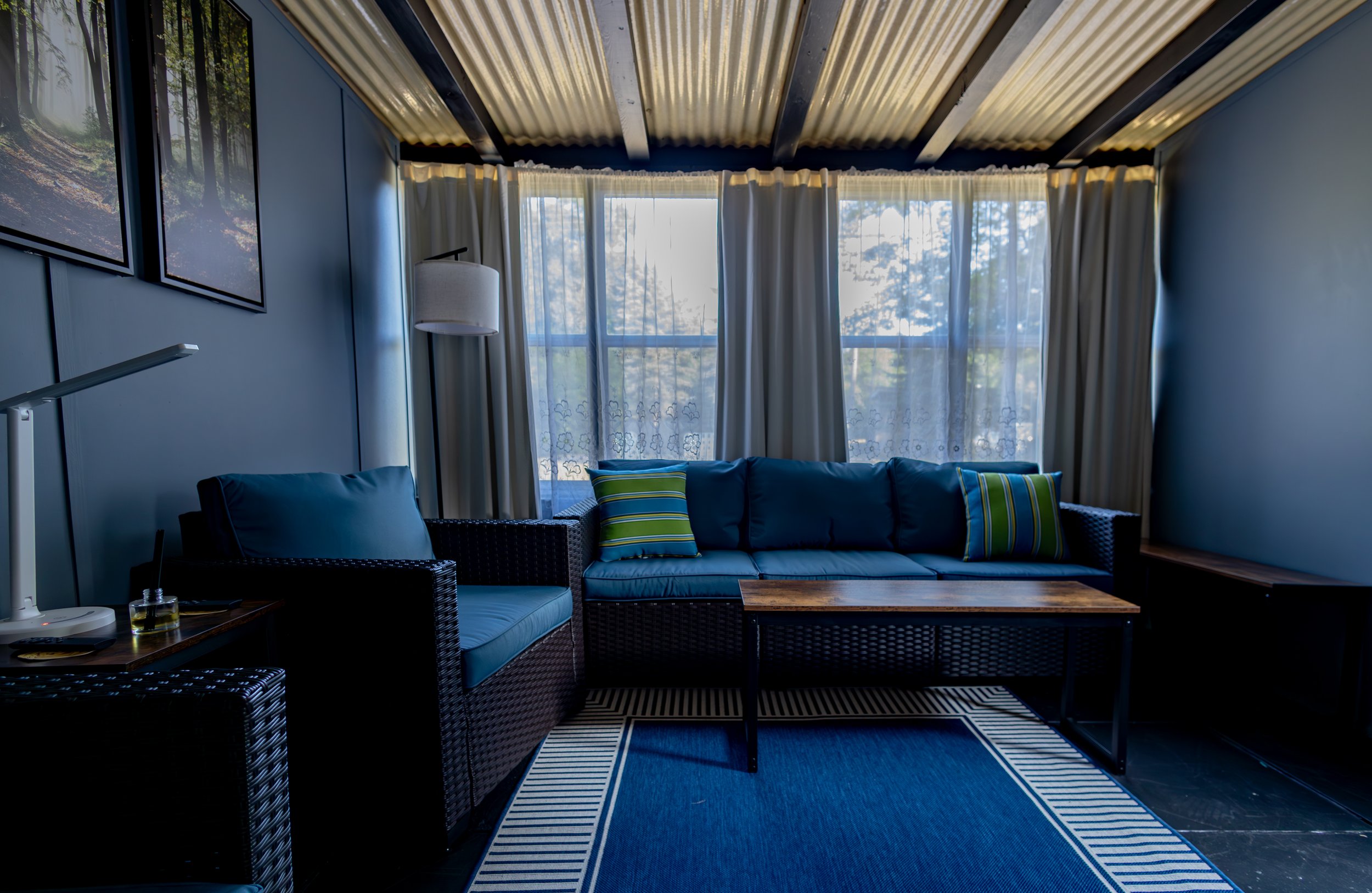 Sun room with blue walls, a dark wicker sofa with blue cushions and striped pillows, a coffee table, a floor lamp, and framed pictures on the wall at The Dunes, with large windows and white curtains letting in natural light.