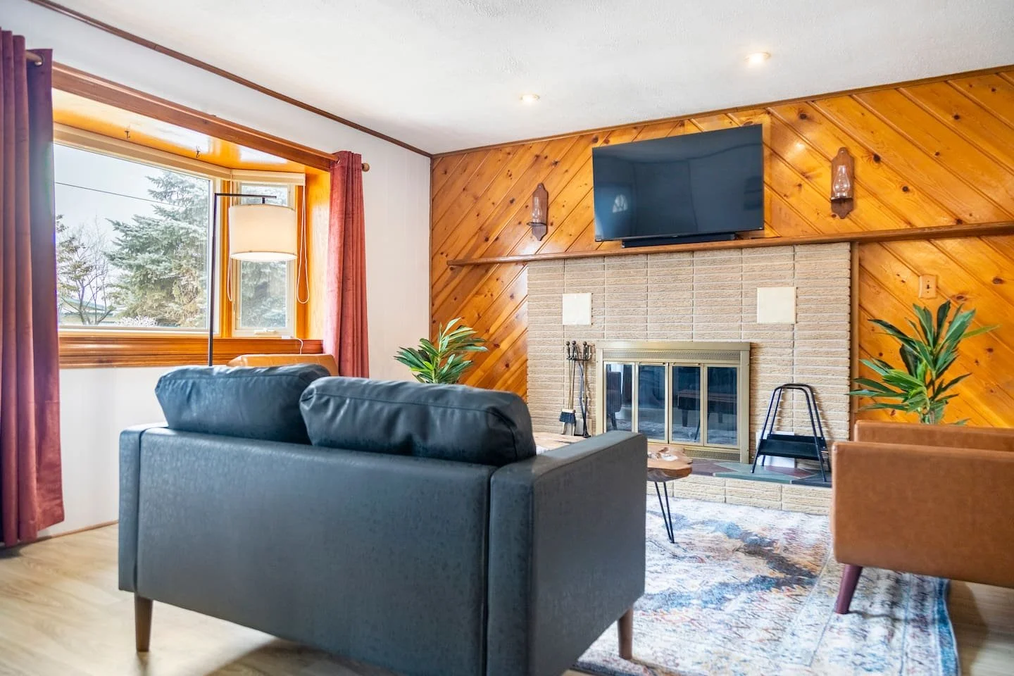 Living room with a black couch, green plants, a large window with pink curtains, a fireplace with a mounted TV, and a carpet at The Canal House