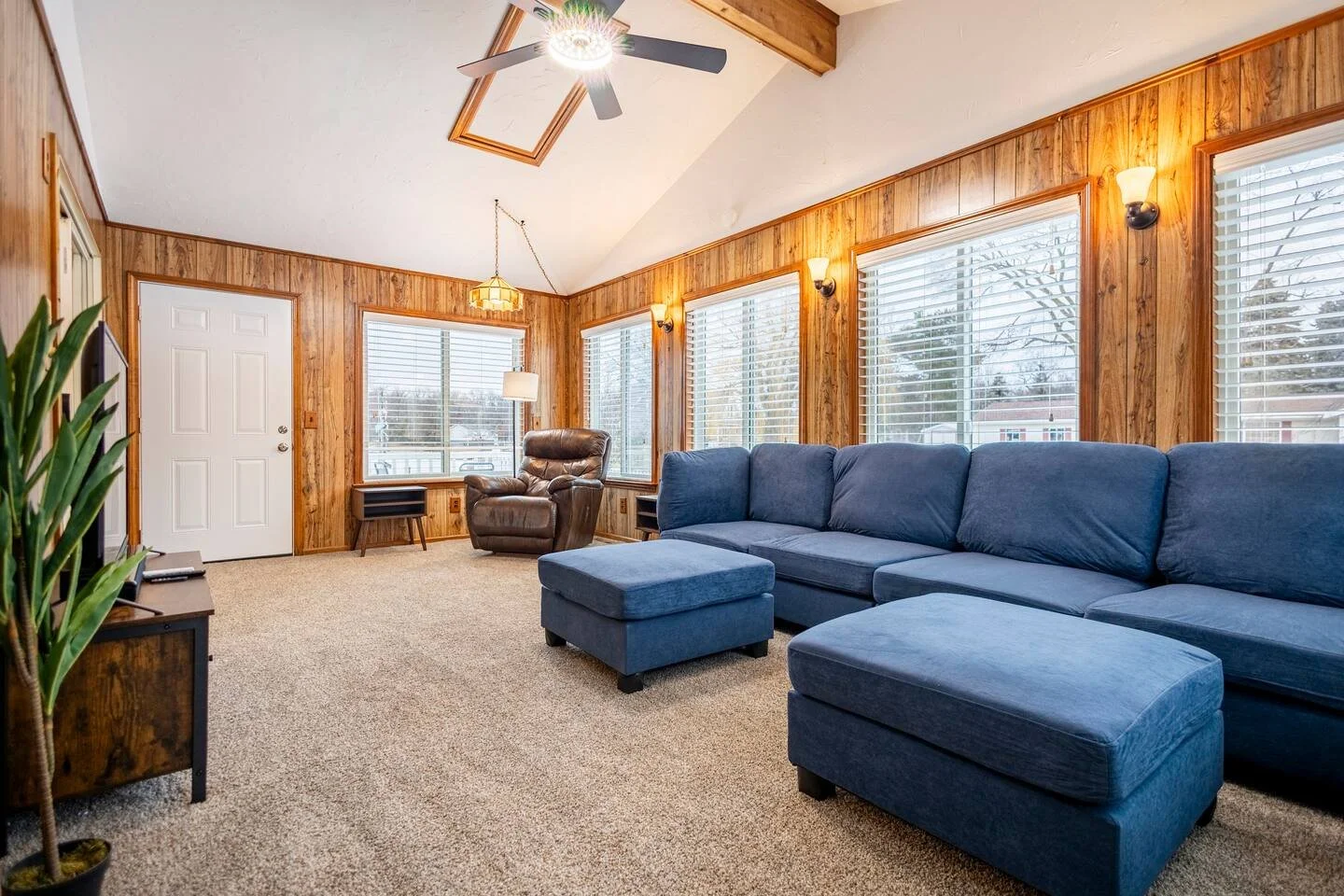 Living room with wood-paneled walls, large windows, a blue sectional sofa, a brown recliner, a ceiling fan, and carpeted floor.