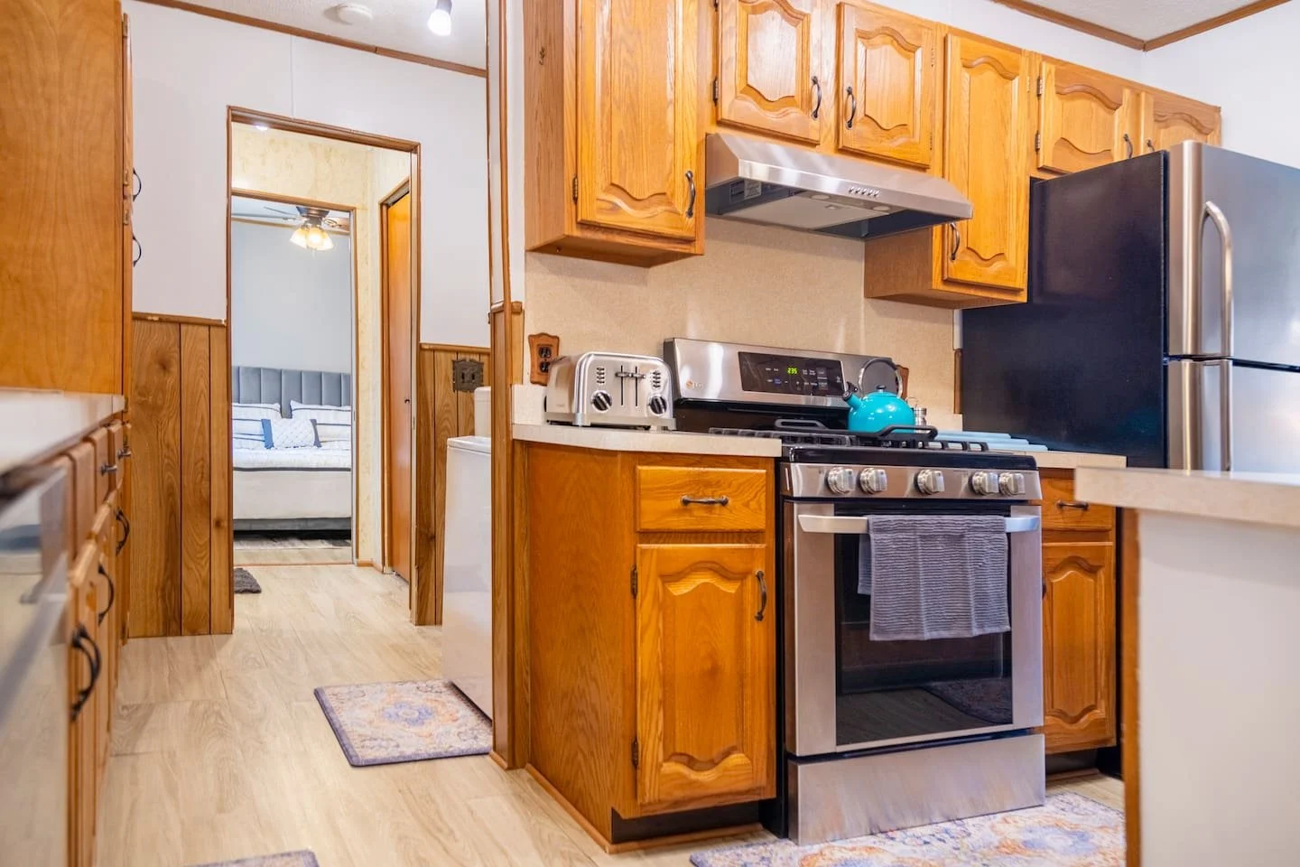 View of a kitchen with wooden cabinets, a stainless steel stove with a blue kettle, a microwave, a refrigerator, and a doorway leading to a bedroom with a bed and nightstand.