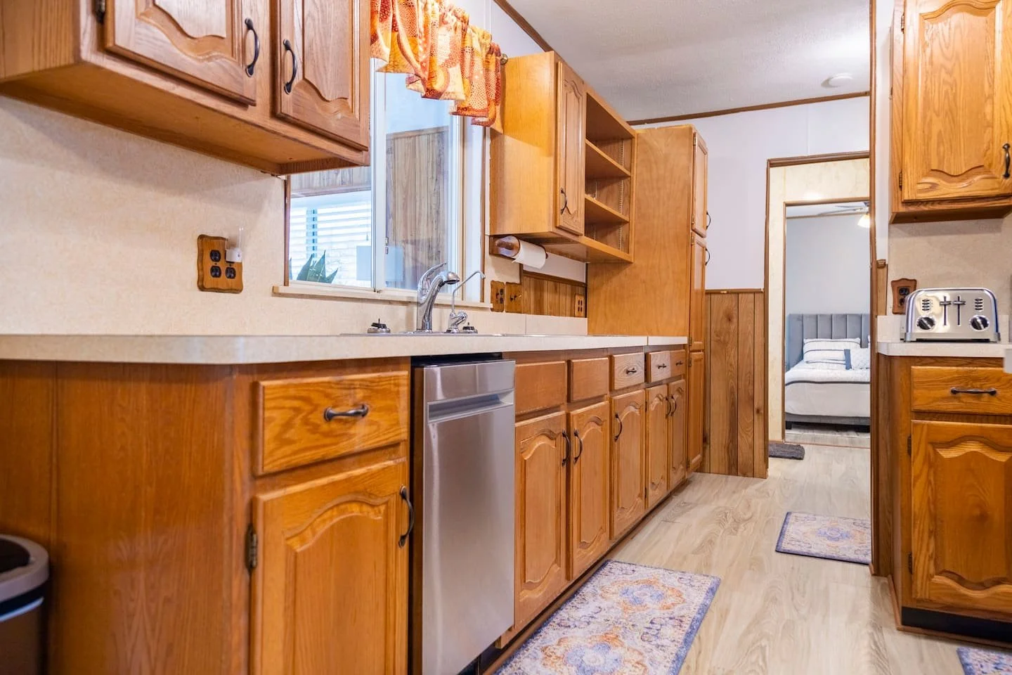 Kitchen with wooden cabinets, a window over the sink, and a glimpse into a bedroom with a bed.