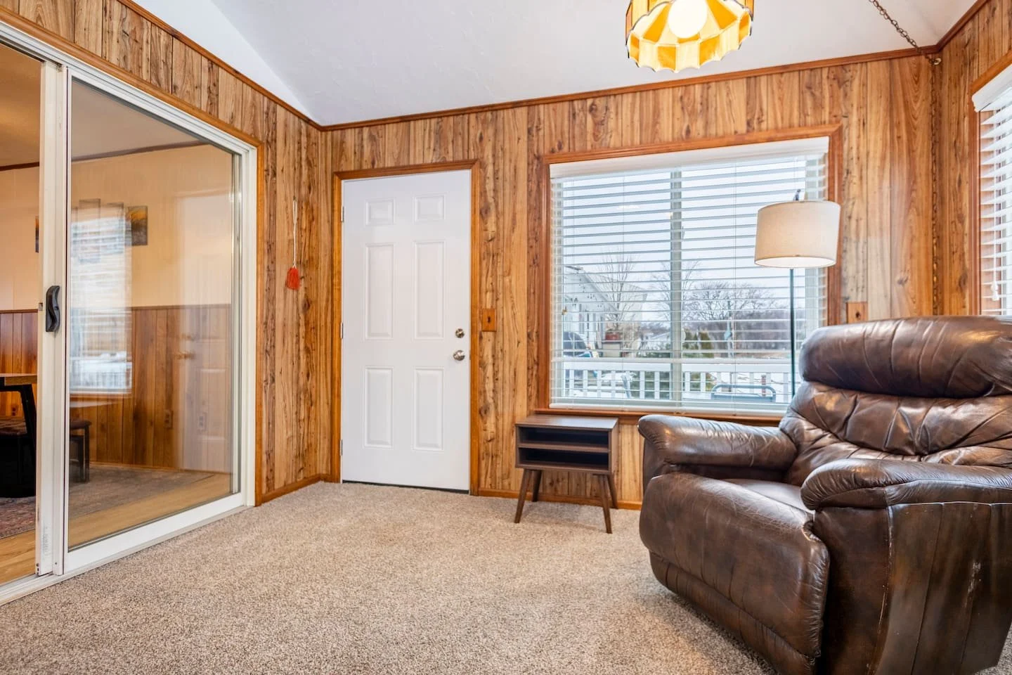 Living room with wood-paneled walls, large windows with blinds, a brown leather armchair, a small wooden side table, a floor lamp, a sliding glass door, and a white front door.