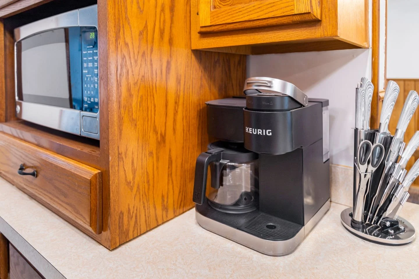 Keurig coffee maker on kitchen countertop next to a utensil holder filled with knives, with a wooden cabinet and microwave in the background.
