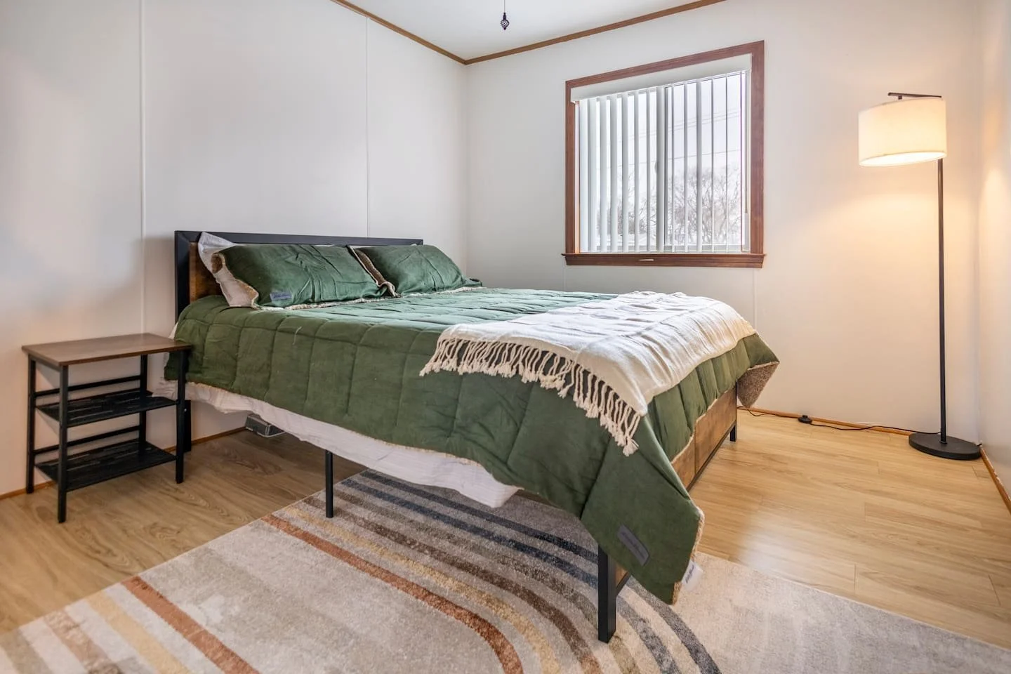 A bedroom with a bed covered with a green quilt and matching pillows, a wooden nightstand, a window with vertical blinds, a standing lamp, a beige and brown striped rug, and hardwood floors.