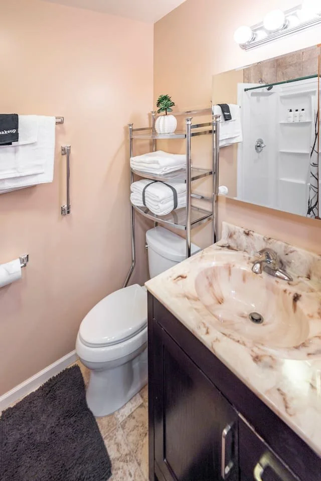 A small bathroom with a beige wall, a white toilet, a marble countertop sink, a mirror, and shelving with folded towels and a small plant. There are towel bars with white towels, and a shower in the background.