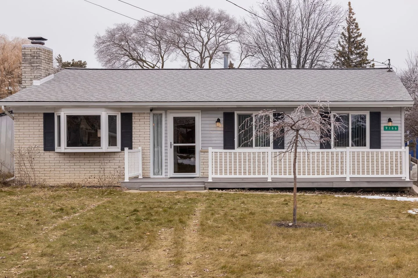 Single-story house with a brick and siding exterior, black shutters, a small porch with white railing, a deck, a leafless tree in front yard, and cloudy sky.