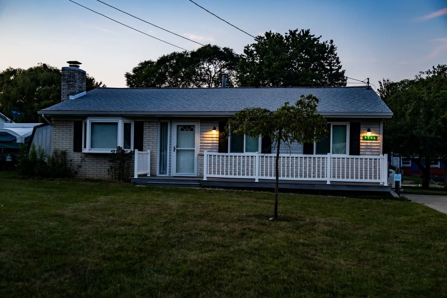 Front view of a small house with a porch, white railing, and a tree in the yard at dusk.