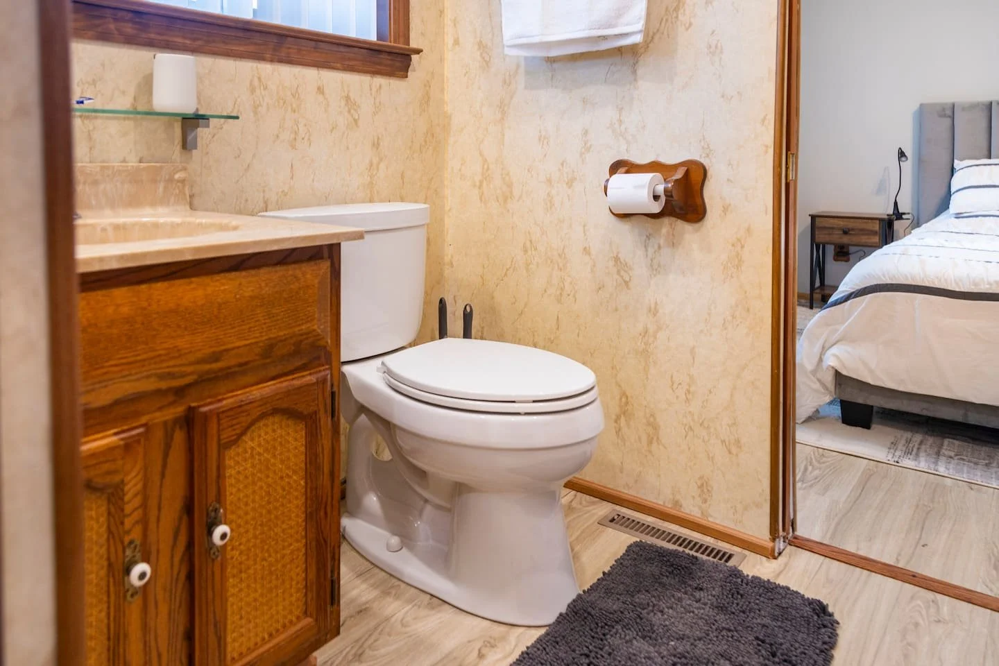 A small bathroom with a toilet, wooden cabinet, and a mirror with a shelf. The bathroom has beige walls, wooden trim, and light-colored flooring. A window with white curtains is visible, and part of a bedroom with a bed and nightstand is seen through