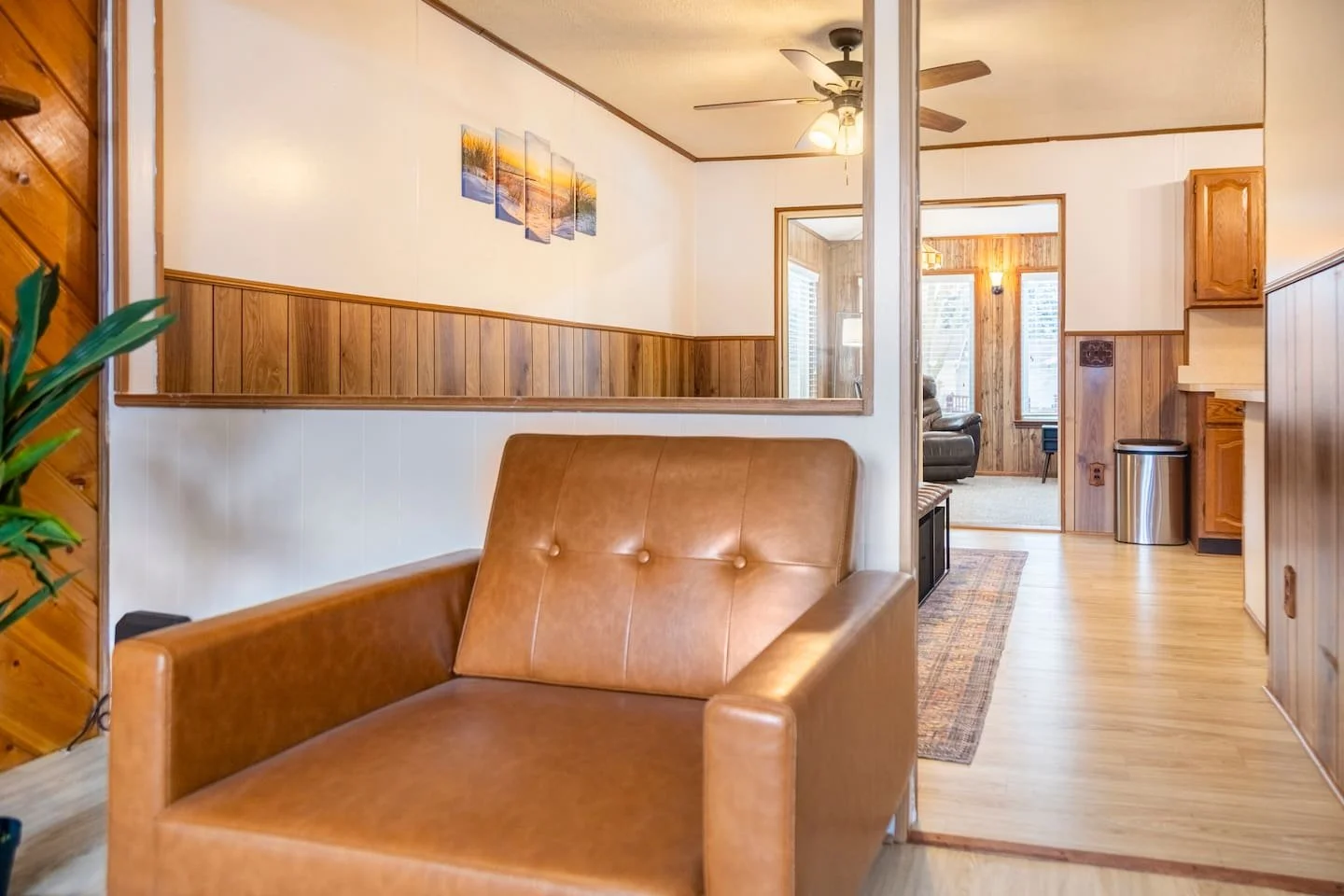 Living room with a brown leather armchair in the foreground, wooden wall paneling, a ceiling fan, and some pictures on the wall; opens into a second room with wooden walls, windows, and furniture.