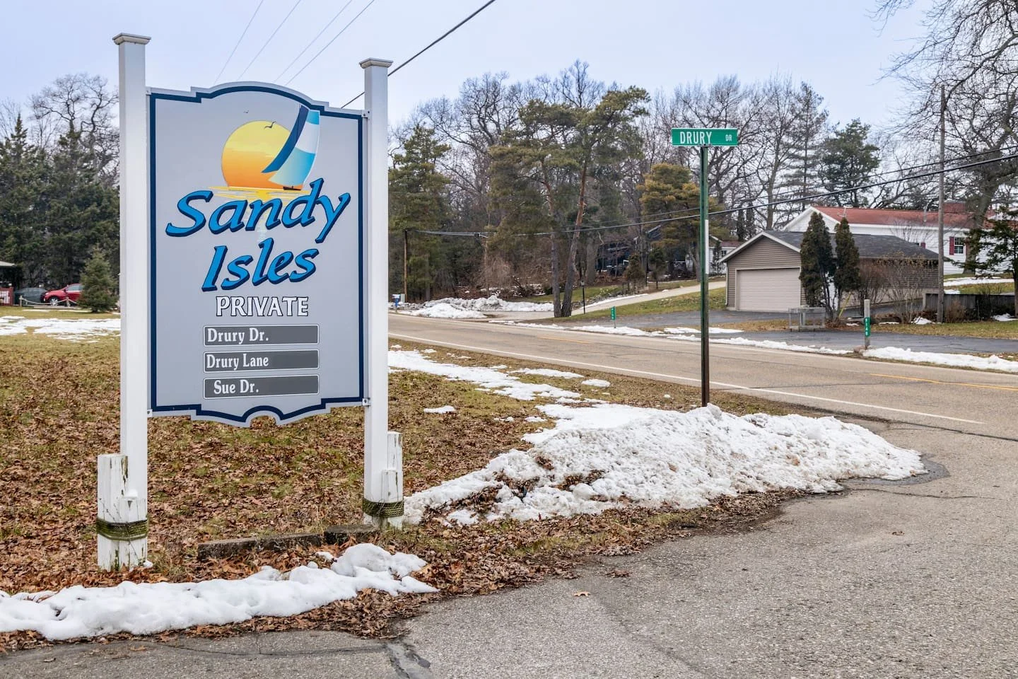 Sign for Sandy Isles private neighborhood with street names Drury Dr, Drury Lane, and Sue Dr. Snow on the ground, houses and trees in the background on a winter day.