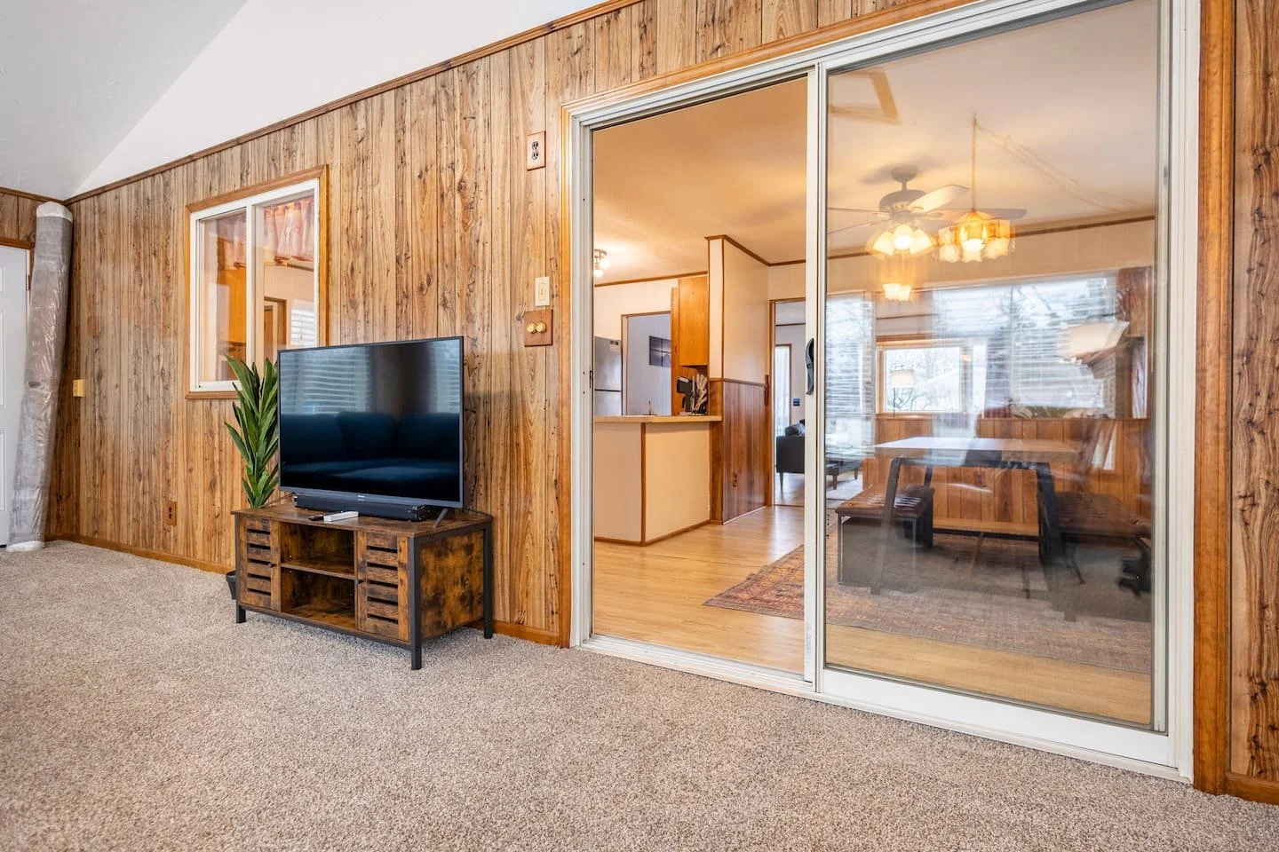 Living room with wood-paneled walls, a flat-screen TV on a wooden stand, and a sliding glass door leading to a dining area with a ceiling fan.