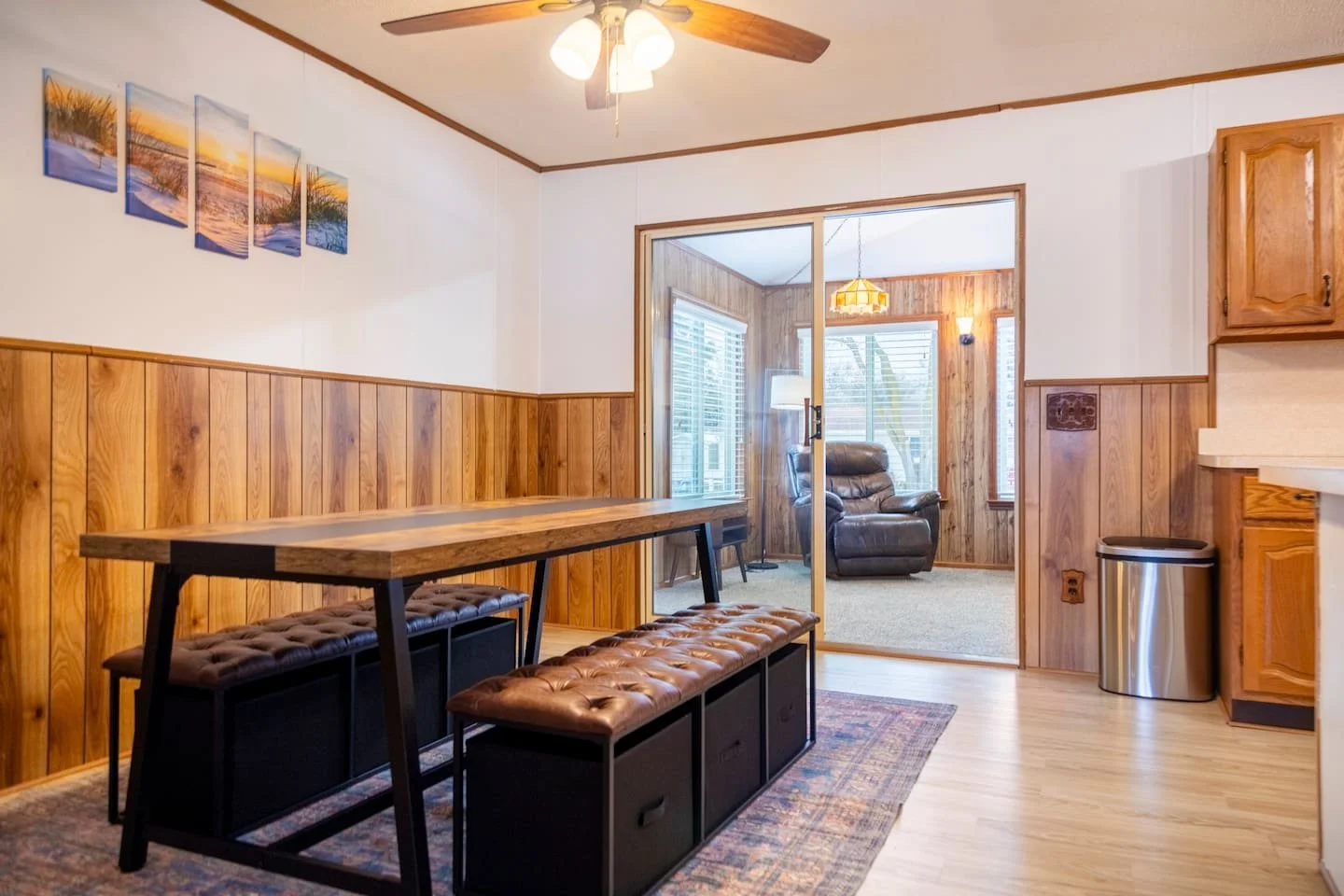 A dining room with wood-paneled walls, a wooden dining table with leather cushioned benches, a ceiling fan with lights, and a view into a sunroom with a recliner, windows, and a hanging lamp.