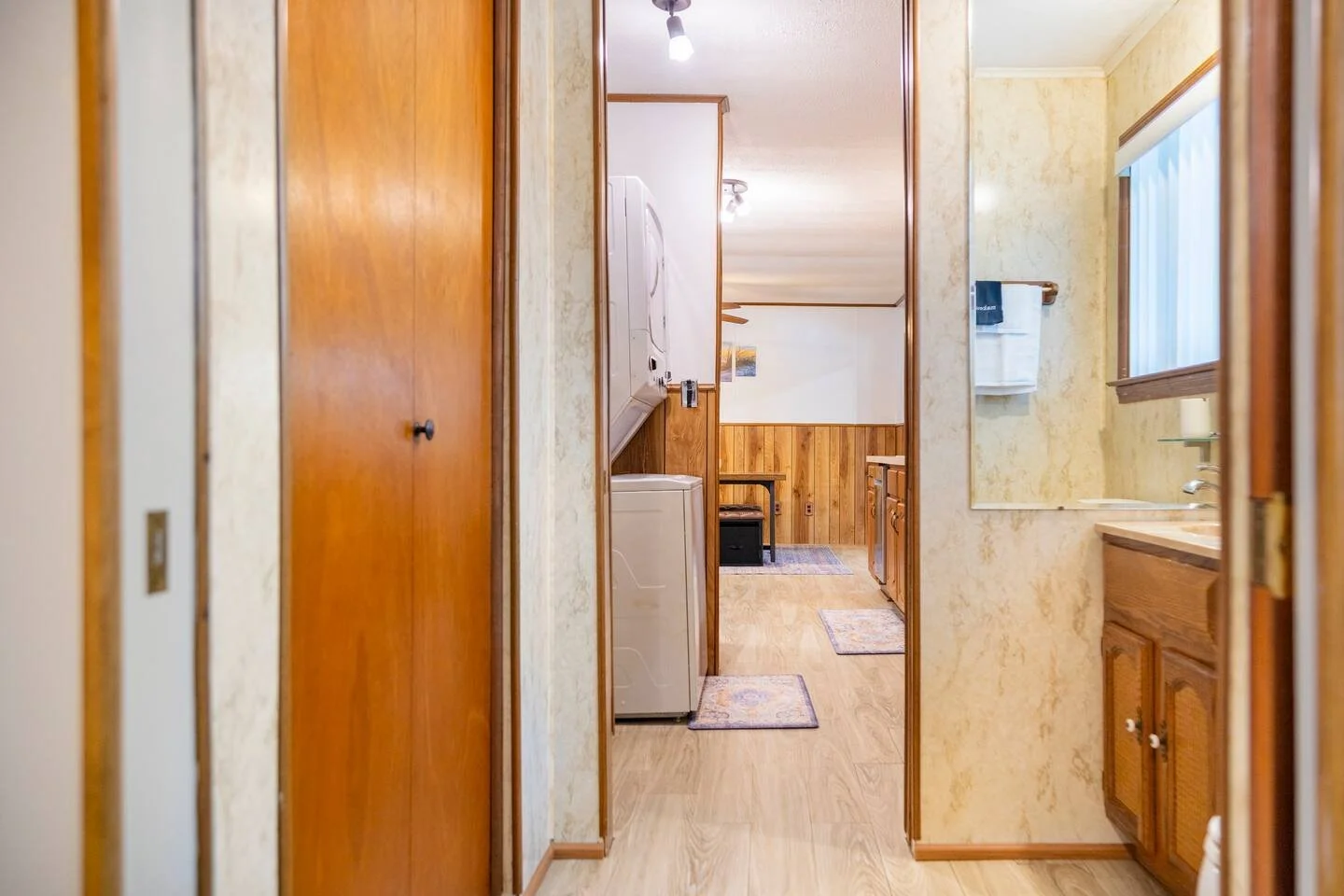 View through a hallway to a laundry area with a washing machine, and a kitchen with wooden cabinets and a window with blinds.