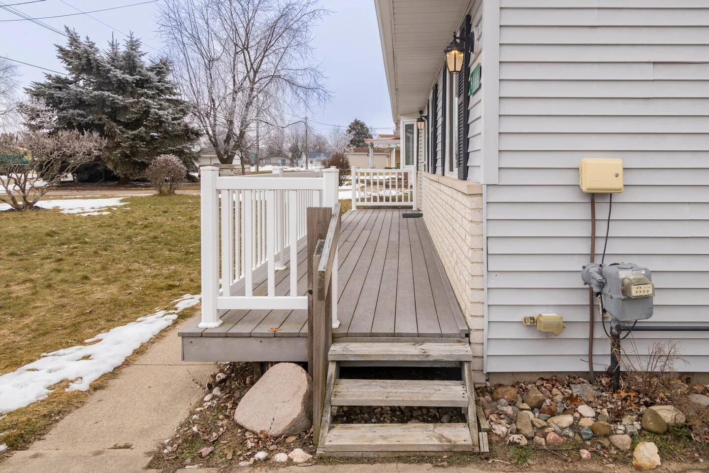 View of a house's screened porch deck with wooden stairs, white railing, and attached utilities, adjacent to a yard with patches of snow and trees in the background.