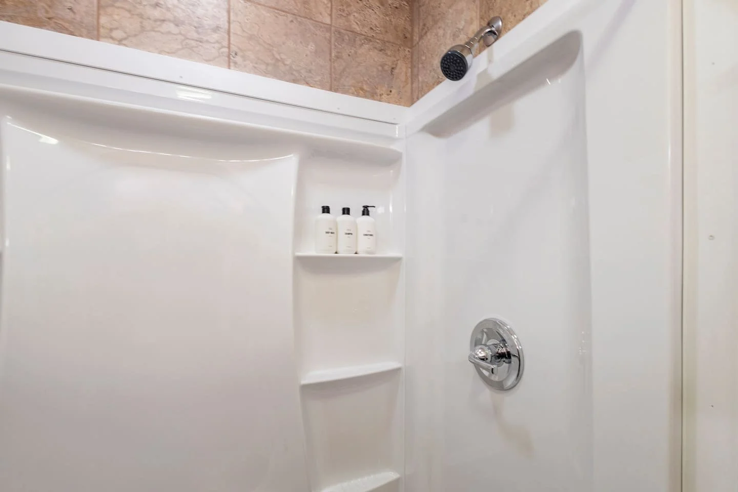 Close-up of a corner shower with built-in shelves holding three white bottles, a silver showerhead, and a round silver control knob on a white wall.