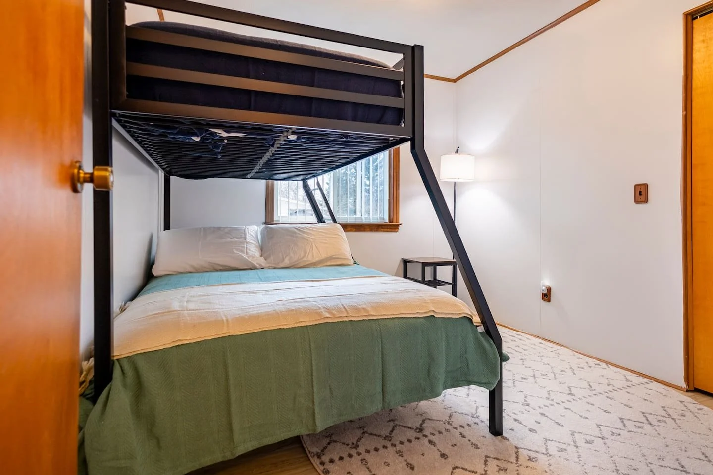 A bedroom with a metal bunk bed, a window, a bed with white pillows and a green bedspread, a small black side table, a floor lamp, and a patterned rug.