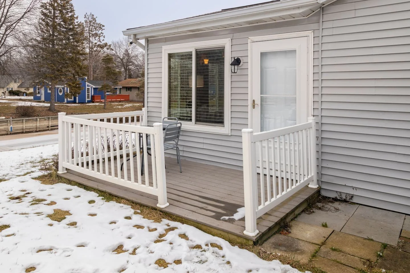 Back patio with white railing, gray wooden deck, small gray table, and two gray chairs, attached to a light gray house with white siding, large window, both door and window with white frames, outside winter scene with snow on the ground, neighboring 