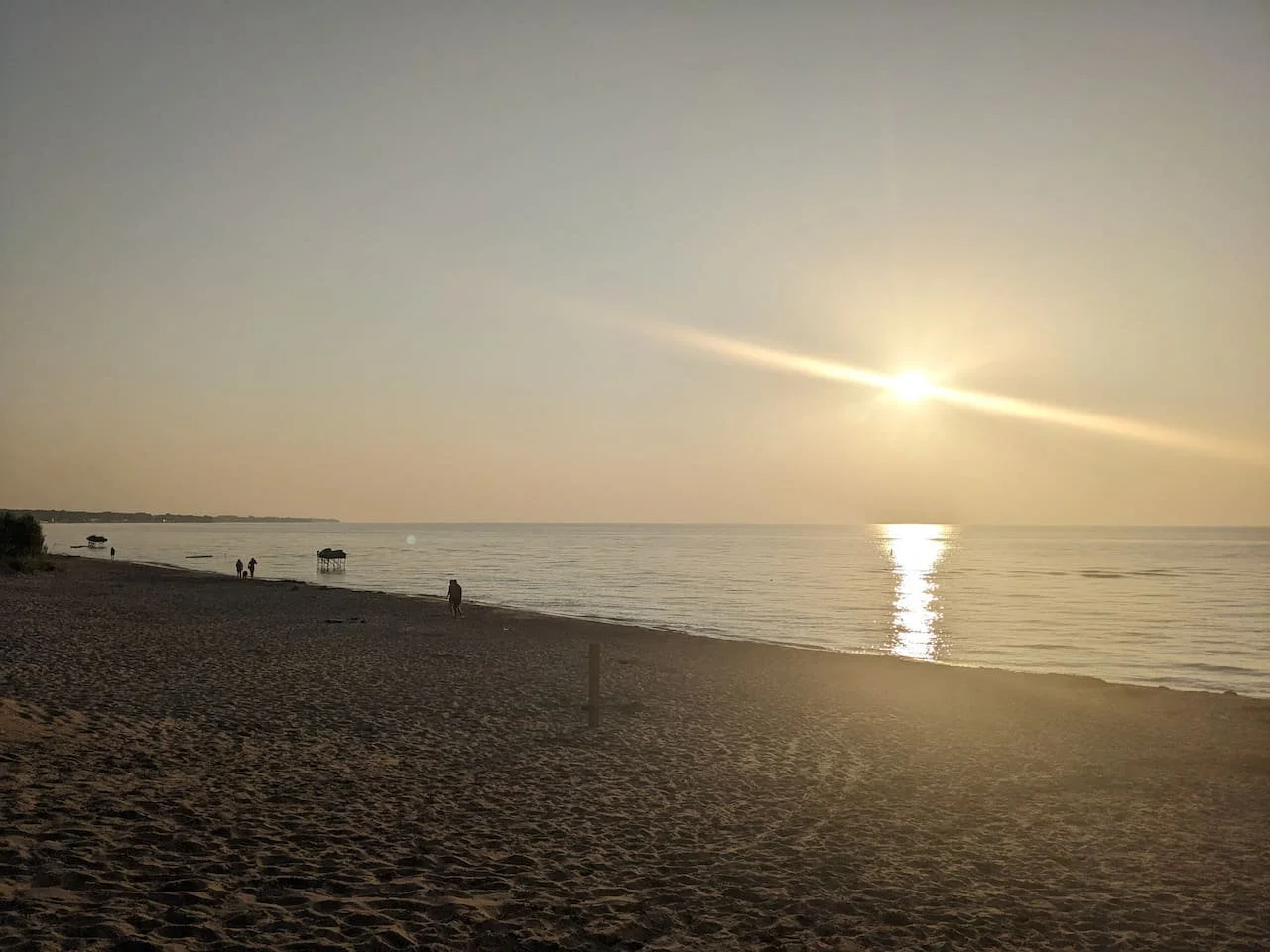 Beach at sunset with a few people walking and boats on the water.