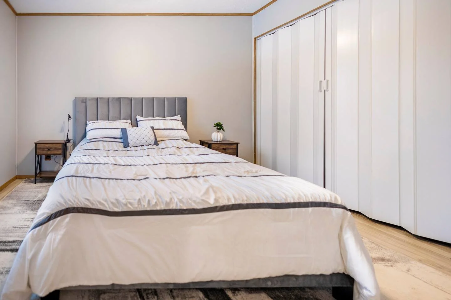 A neatly made bed with a gray upholstered headboard, white and gray bedding, and decorative pillows, in a minimalist bedroom with a nightstand with a small plant, a window with white blinds, and a closet with white sliding doors.