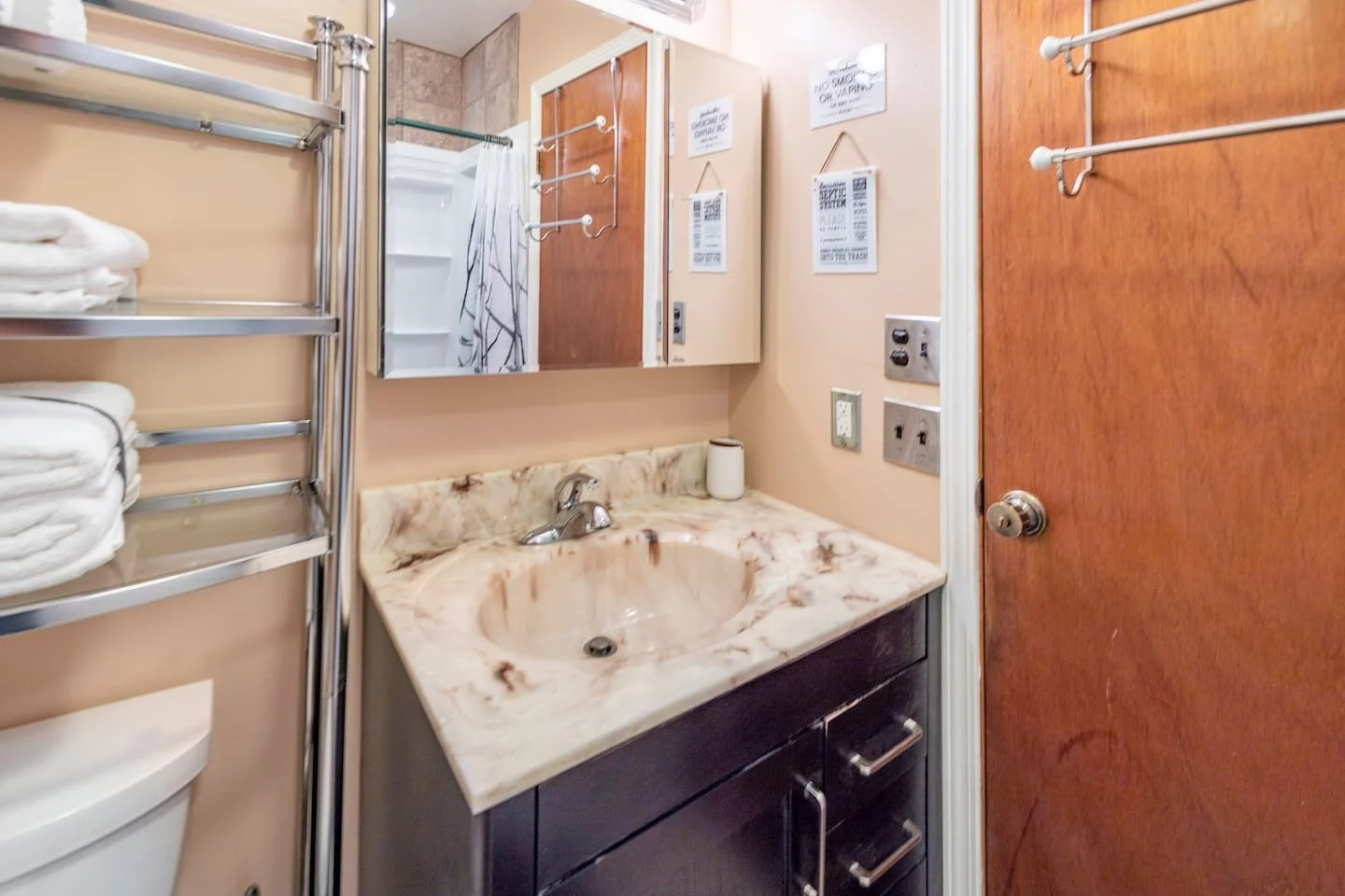 A small bathroom with a marble countertop sink, dark wood cabinet, wall mirror, towel rack, and various signs and electrical outlets on beige walls.