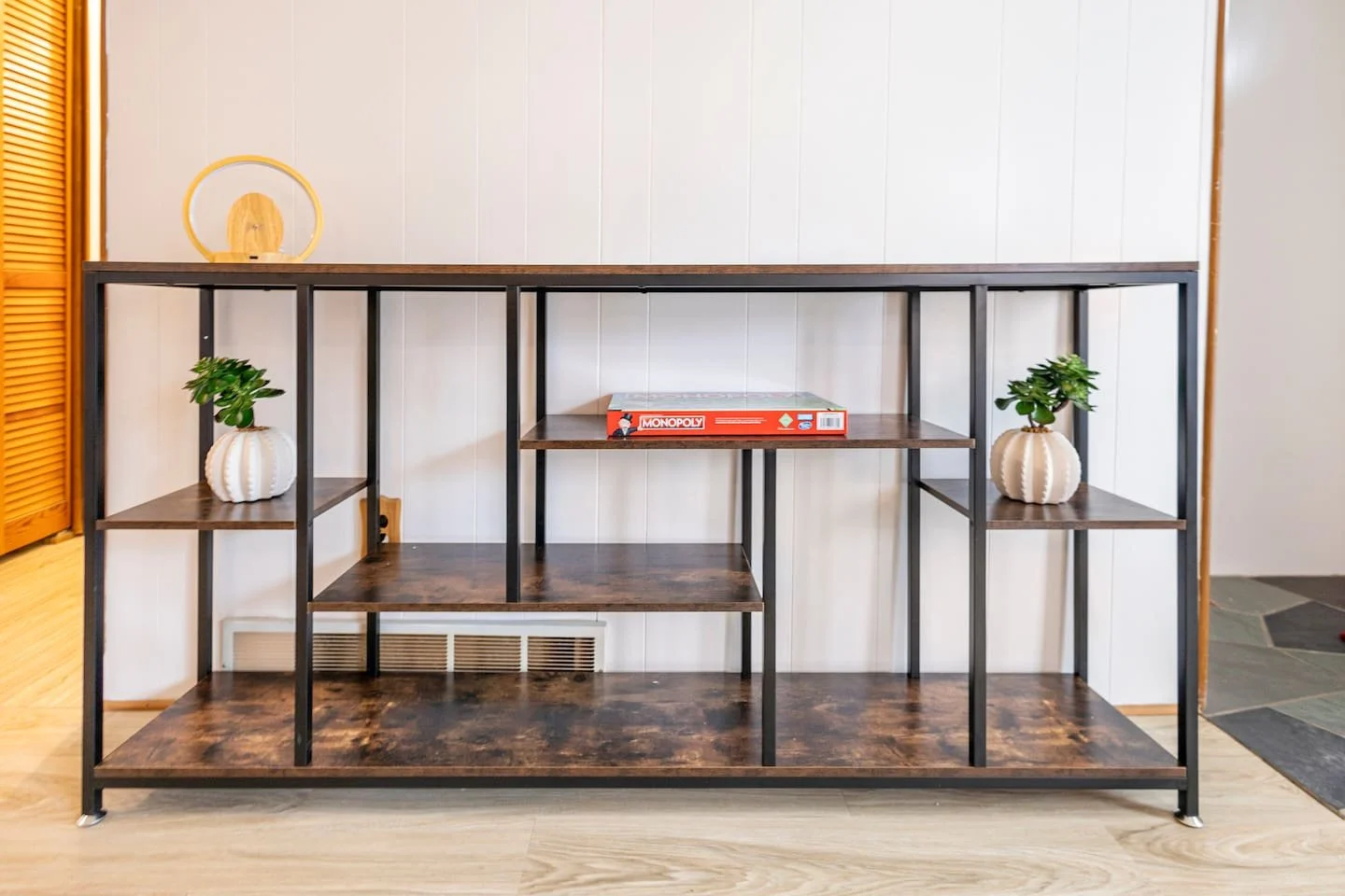 Empty wooden and metal shelving unit with three potted plants and a Monopoly board game on the middle shelf, against a white paneled wall.