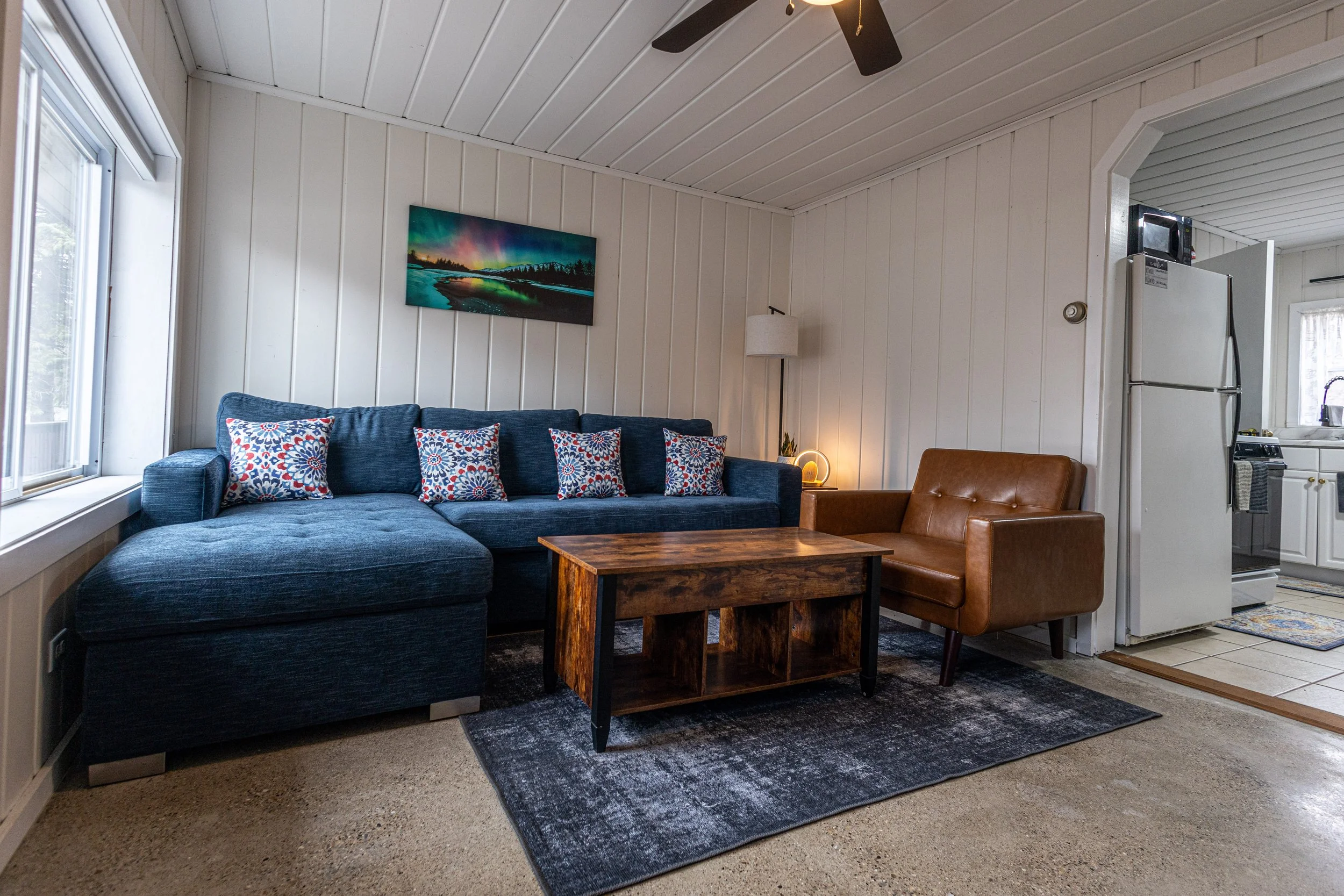 Living room with a blue sectional sofa, patterned pillows, a brown leather chair, a wooden coffee table, a floor lamp, and a wall art of aurora borealis over a lake. The room has white paneled walls and is adjacent to the kitchen with visible refrigerator and stove.