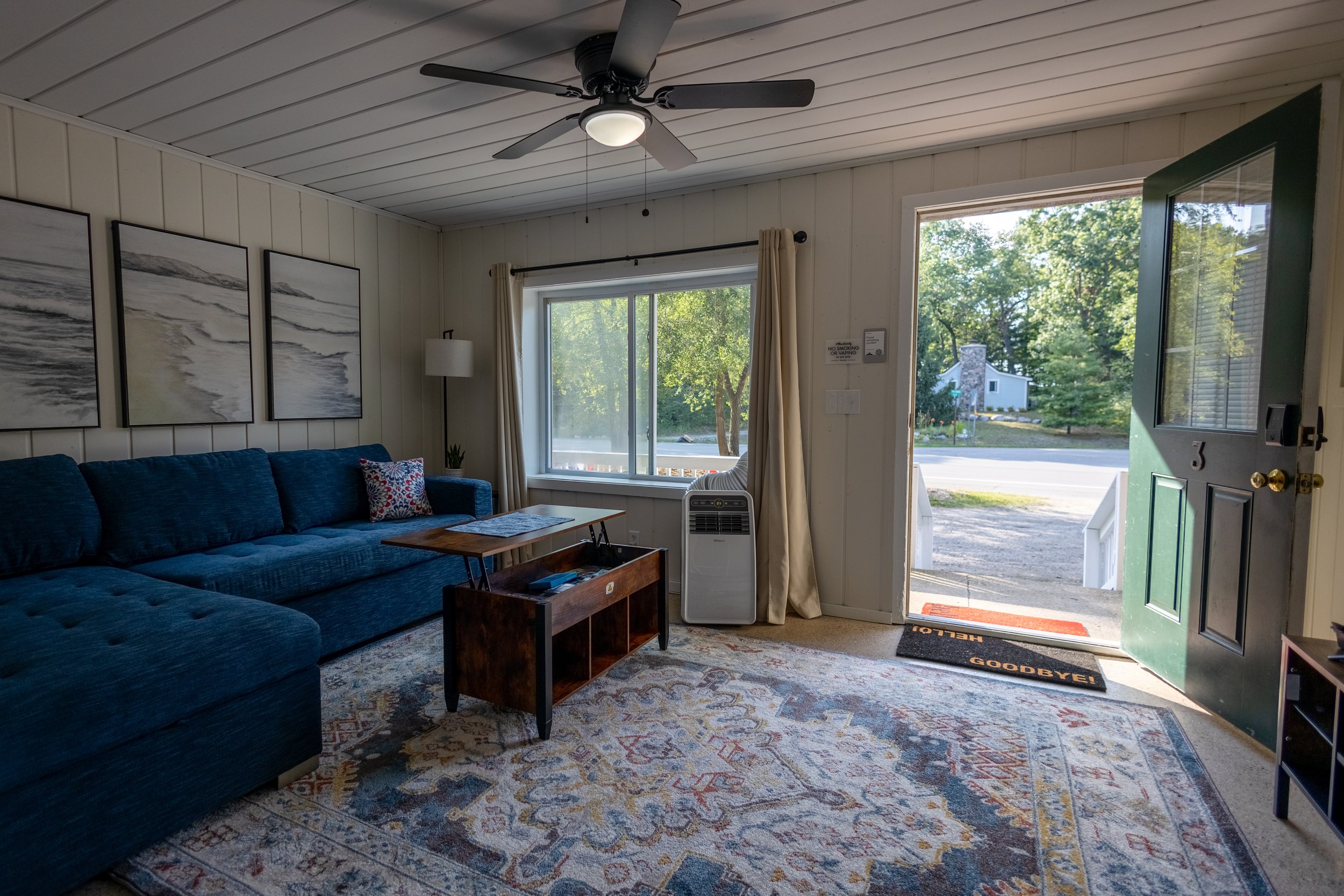 Living room with a navy blue sectional sofa, framed abstract art on white paneled walls, a wooden coffee table, an air conditioning unit, a large window with beige curtains, a ceiling fan, and an open green front door with a welcome mat outside.