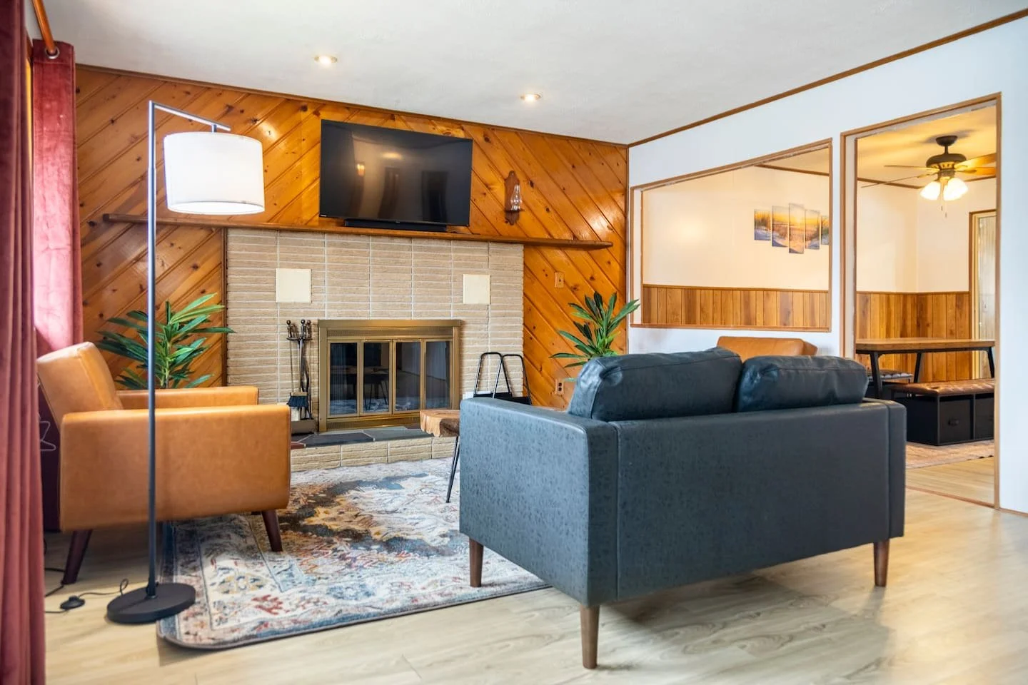Living room with a fireplace, mounted flat-screen TV, armchair, loveseat, and a patterned rug. There's wooden paneling on the wall behind the fireplace and a mirror reflecting another room with wood-paneled walls and ceiling fan.