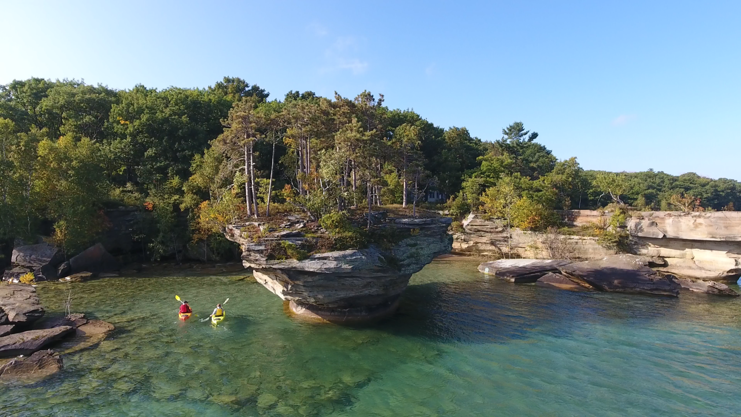 Two people kayaking on a river with Turnip Rock iconic formation and green trees under a clear blue sky in Port Austin, Michigan
