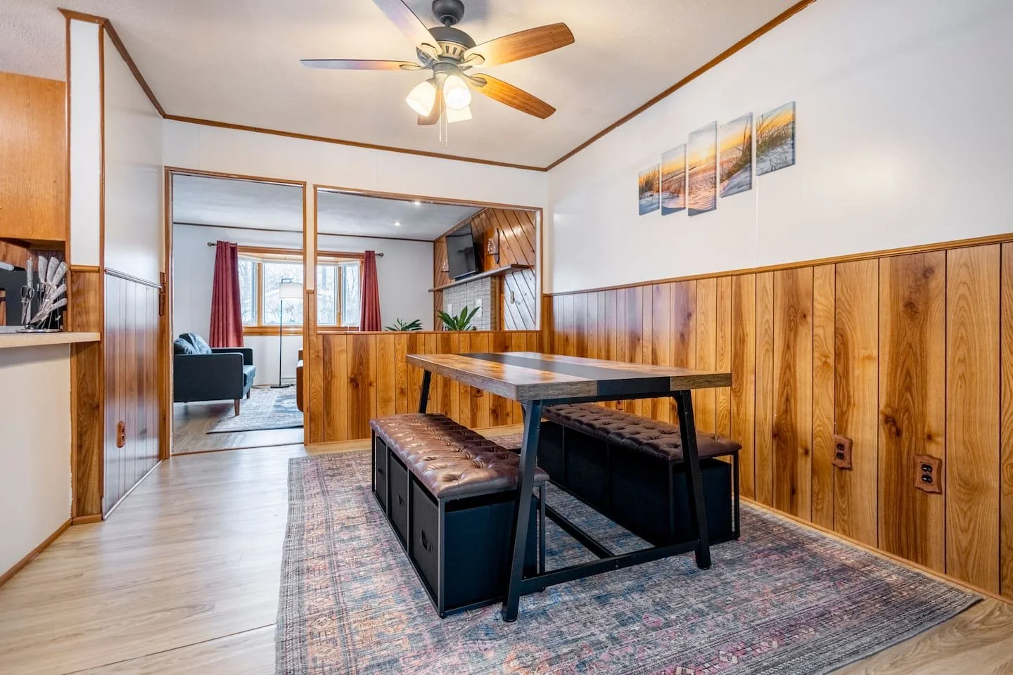 Dining area with a wooden table, brown cushioned benches, wood-paneled walls, and a ceiling fan, with a view into the living room with red curtains and a fireplace.