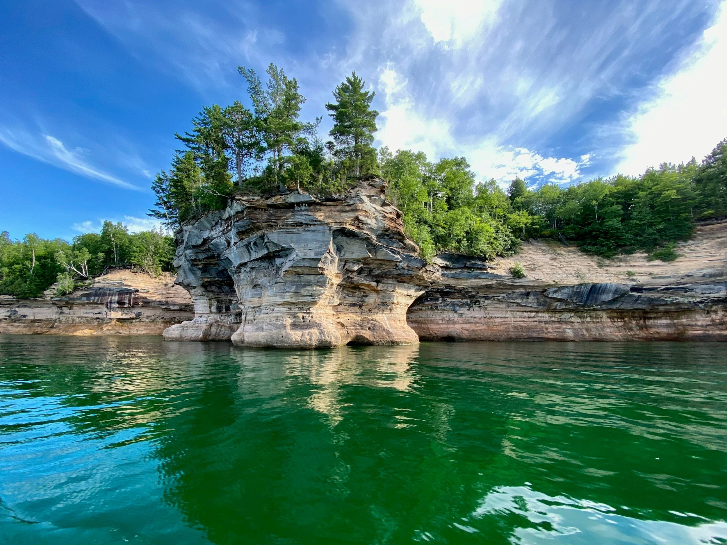 A scenic view of a rocky shoreline with layered cliffs, green trees on top, and a clear blue sky reflected in calm green water.