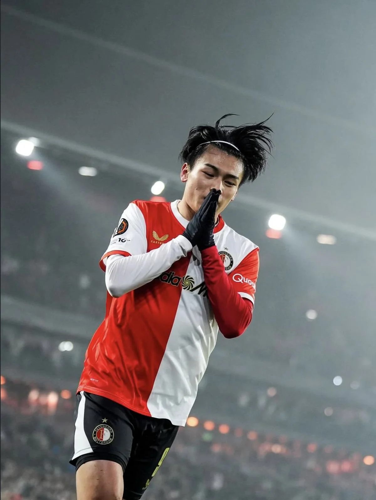A soccer player wearing a red and white jersey, black shorts, and black gloves, standing on a soccer field with stadium lights in the background, holding his hands together in a praying gesture.