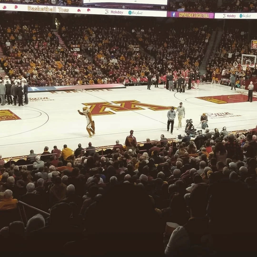 Basketball court at a game with a mascot walking near the stands, surrounded by spectators. The court has the Minnesota Golden Gophers logo at center, and there are teams and officials on the court.