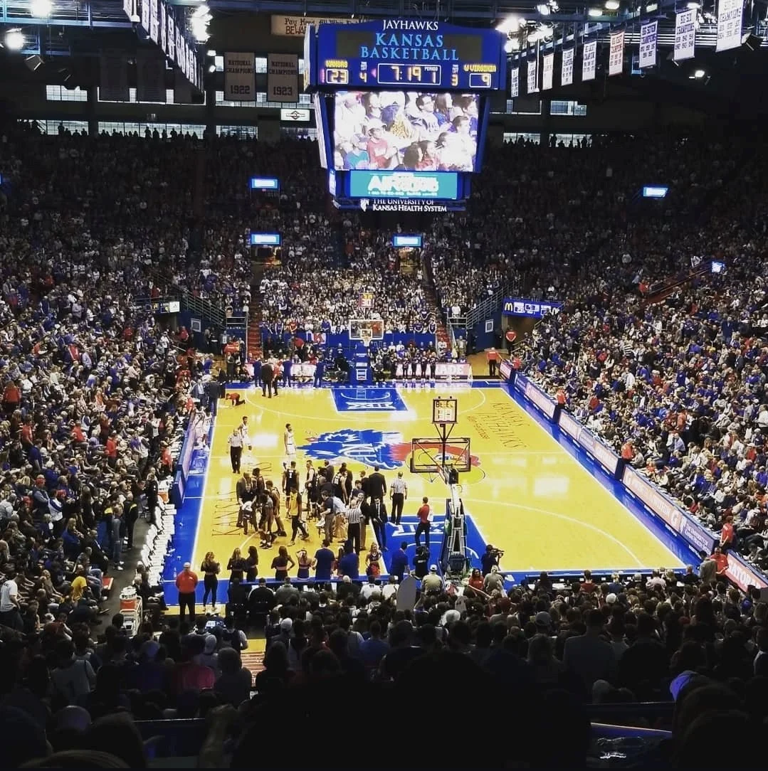 Indoor basketball arena filled with spectators, players on the court, and a large scoreboard showing the Kansas Jayhawks playing against UConn, with about 7 minutes remaining in the game.