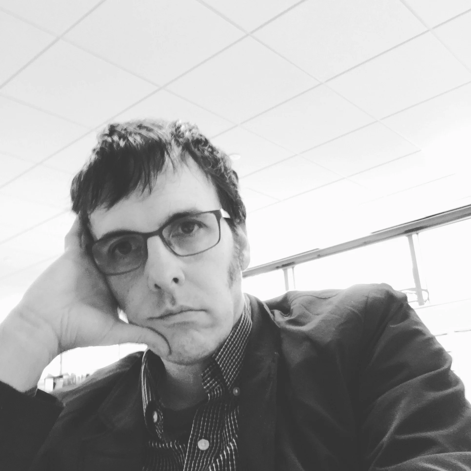 A black-and-white photo of a man with short, dark hair, glasses, and a serious expression, resting his head on his hand in an indoor setting.