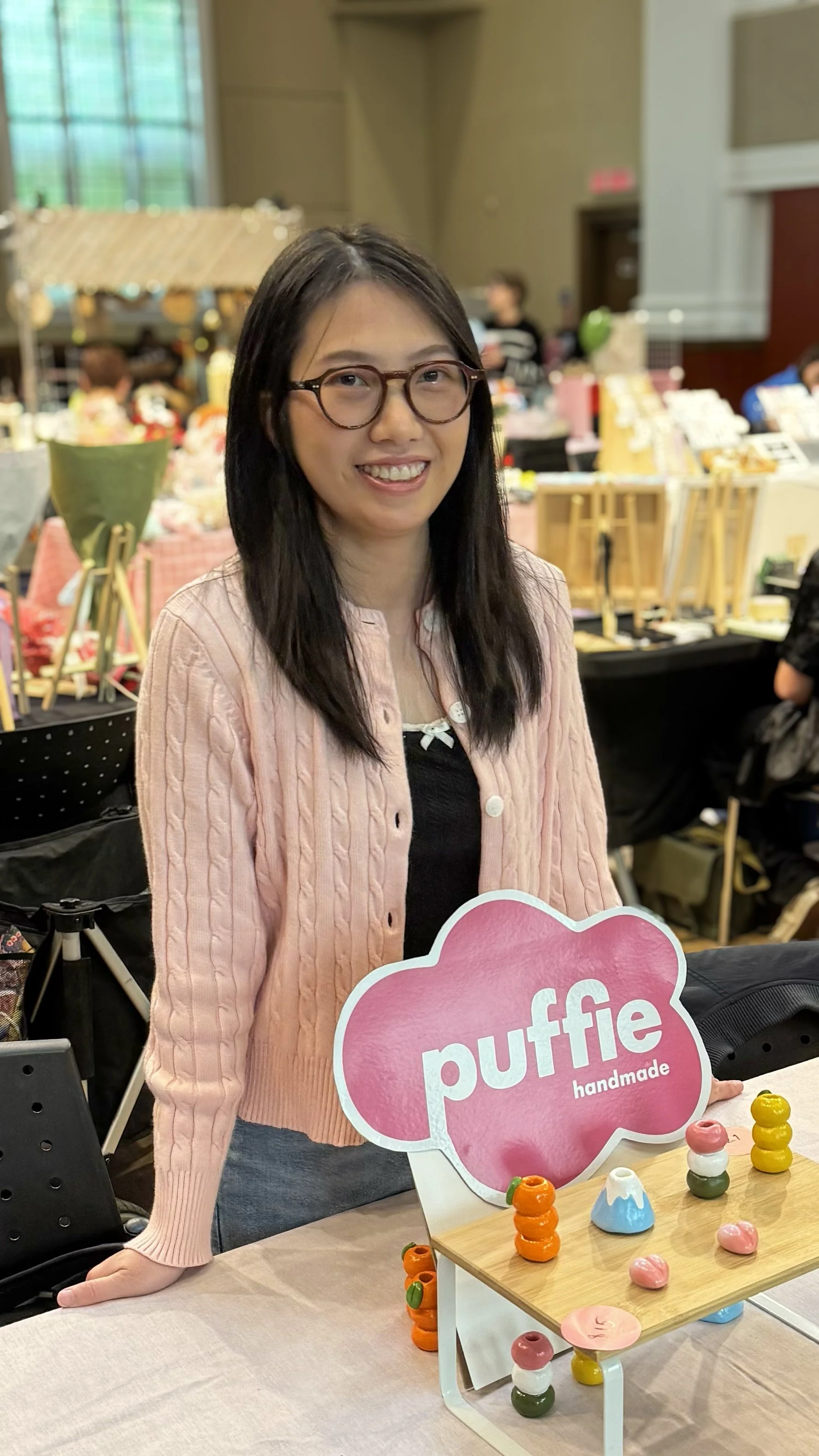 A woman with glasses standing behind a display at a craft fair, with a pink sign that says 'puffie handmade'; colorful small handmade ceramic or clay items are on the table in front of her.
