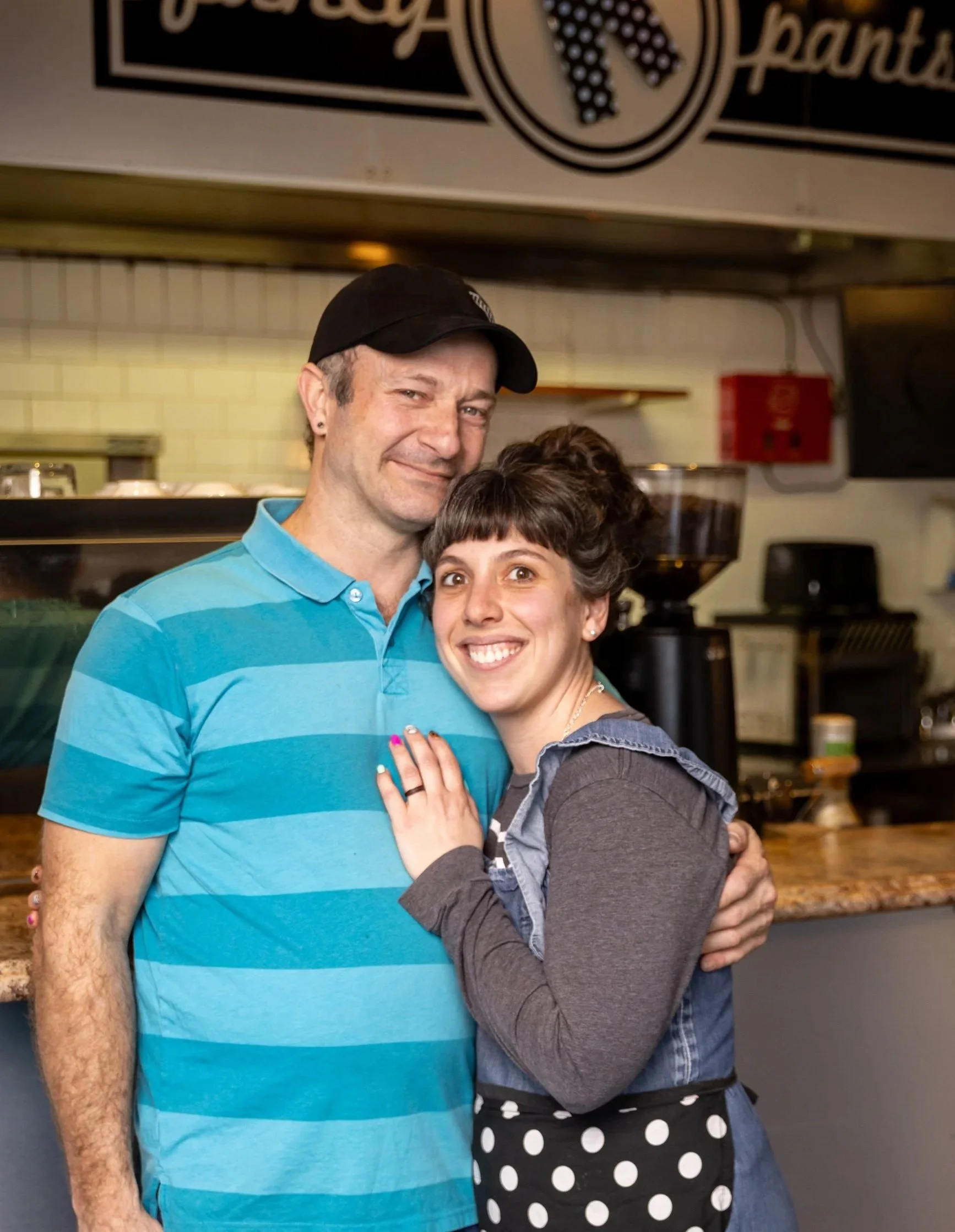 Hayley and Neil Cullington of Fancy Pants Cafe in Bridgewater, NS hug and smile at the camera from in front of their kitchen.